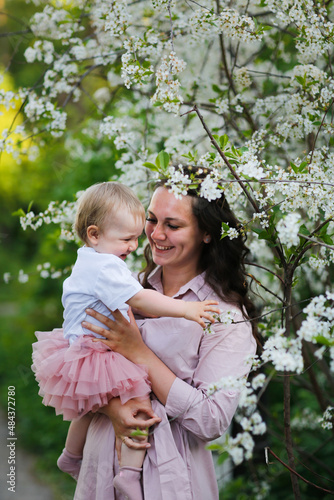 Lovely beautiful feminine mom with her daughter in her arms in spring with cherry blossoms. female natural beauty and beautiful wavy hair. Young European woman in a dress with a baby in a skirt. © natalialeb