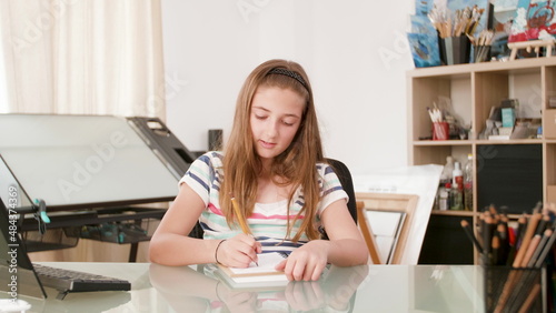 Young schoolgirl writing mathematics exercices on notebook studying for online lesson siting at desk in living room. Schoolkid using elearning schhol platform during coronavirus quarantine