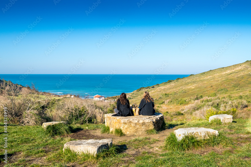 Deux femmes brunes habillées en noir assises sur un rocher sur fond de prairie observant l'océan avec un ciel bleu Deux femmes brunes habillées en noir assises sur un rocher sur fond de prairie observant l'océan avec un ciel bleu