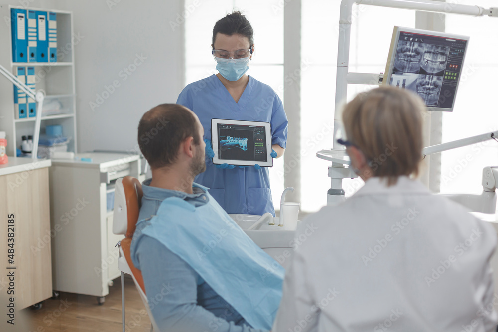 Dentist assistant wearing face mask showing teeth radiography using tablet to patient discussing medical procedure during stomatology examination in dental office. Sick man having oral infection Dentist assistant wearing face mask showing teeth radiography using tablet to patient discussing medical procedure during stomatology examination in dental office. Sick man having oral infection