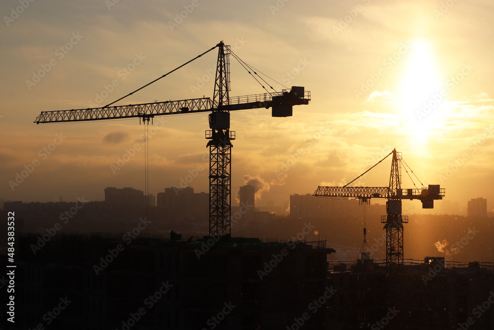 Silhouettes of construction cranes and unfinished residential building during snow on sunrise background. Housing construction, apartment block in winter city during blizzard