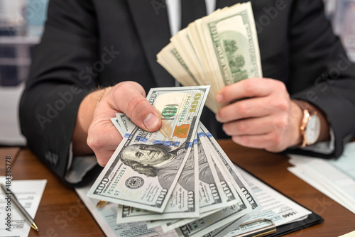  businessman in a black stylish suit at a spacious table counts dollars and fills out forms.