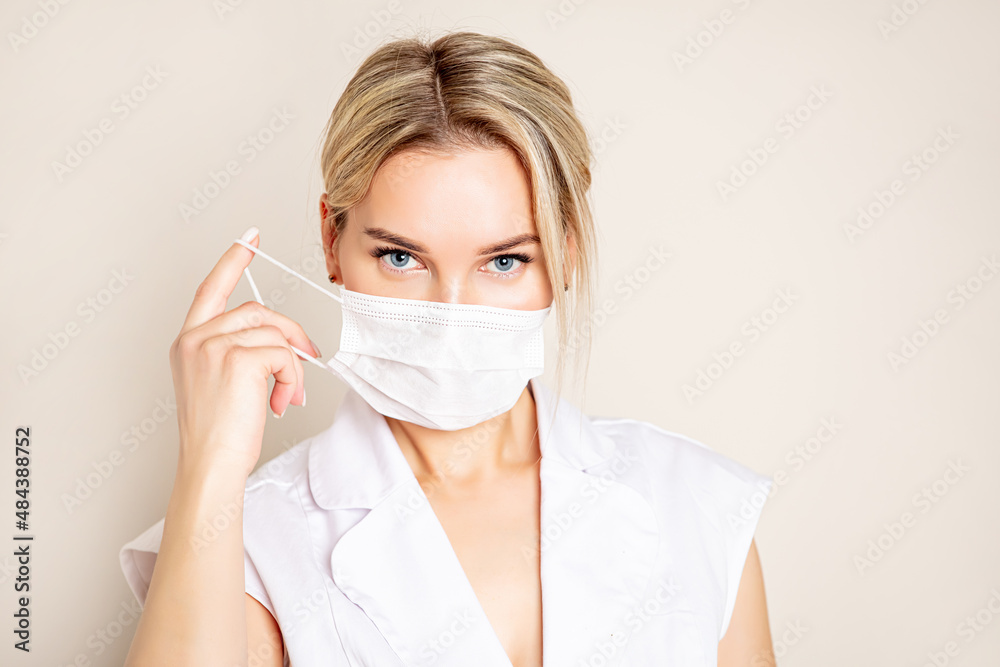 Portrait of a beautiful blonde woman with blue eyes wearing a white medical mask on a light background, looking straight Portrait of a beautiful blonde woman with blue eyes wearing a white medical mask on a light background, looking straight