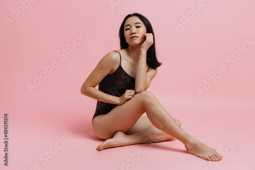 Full-lngth horizontal studio portrait of tender woman sitting and posing in bodysuit isolated over pink background