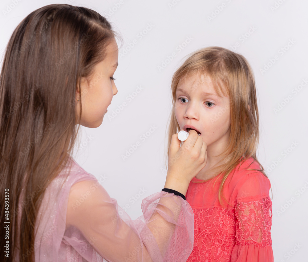 Two girls friends or sister applying lipstick on lips. Helping each other doing make-up to prepare for the holiday. Together having fun isolated over white background Two girls friends or sister applying lipstick on lips. Helping each other doing make-up to prepare for the holiday. Together having fun isolated over white background