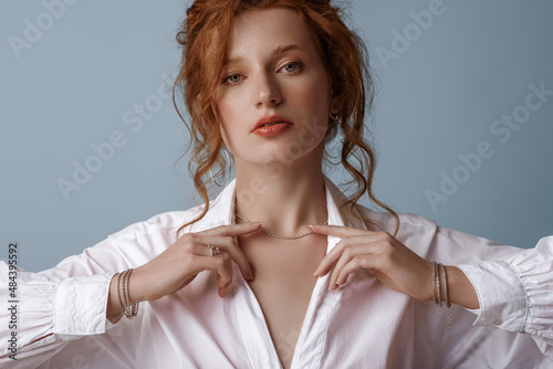 Beautiful elegant redhead freckled woman wearing luxury silver jewelry: earrings, chain, bracelets, white shirt, posing in studio, on blue background. Jewellery advertising concept. Close up portrait