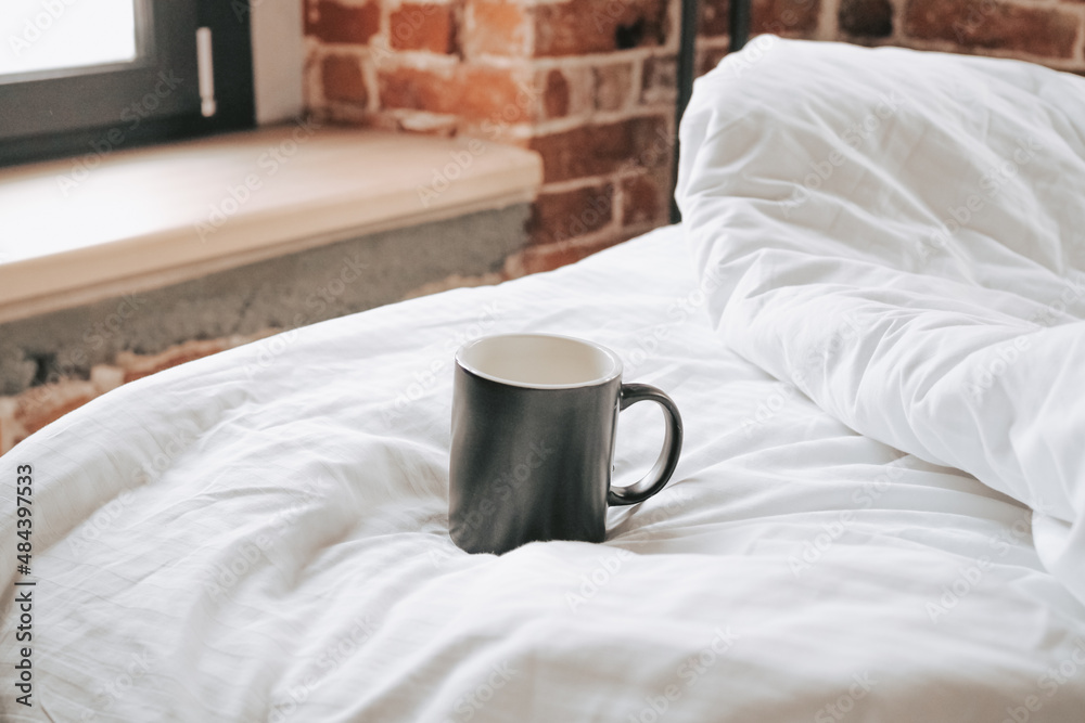 Black mug with hot coffee on a white blanket in the morning. Early wake-up and morning coffee in the hotel bed. Stylish apartments and white bed linen