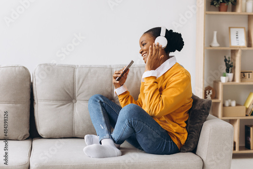 People, technology and leisure concept - happy african american young woman sitting on sofa with smartphone and headphones listening to music at home