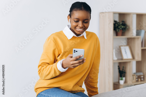 Smiling black girl messaging on smartphone at home, relaxing on comfortable couch, enjoying weekend, copy space