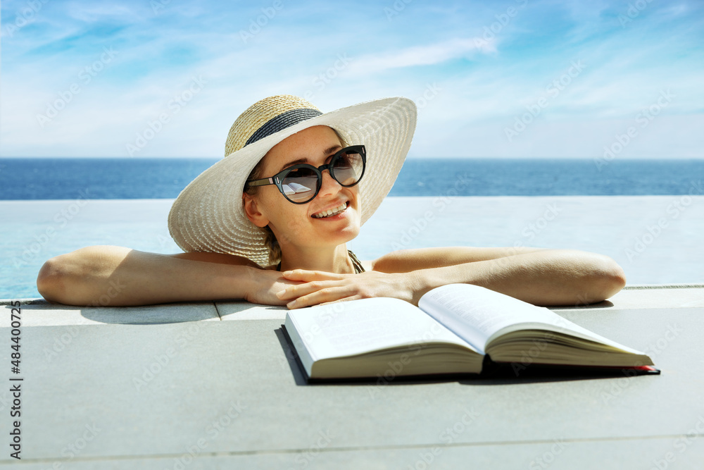 smiling attractive woman with hat and sunglasses reading a book and relaxing in infinity swimming pool at vacation resort