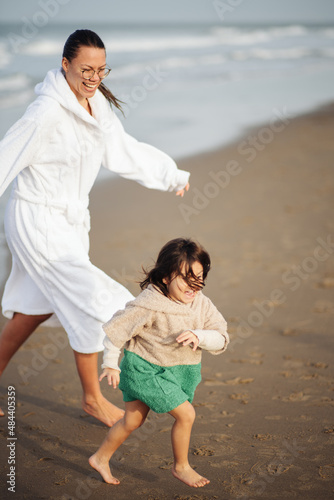 Mother running after a toddler, both with a bathrobe, on the beach during winter in the north sea of the Netherlands 