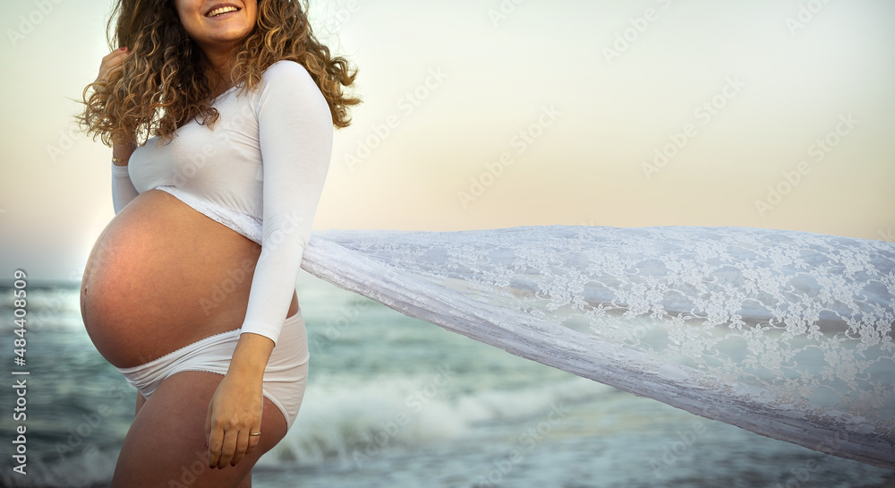 Wind blowing tail of white dress of pregnant latina female smiling by the sea at sunrise Wind blowing tail of white dress of pregnant latina female smiling by the sea at sunrise