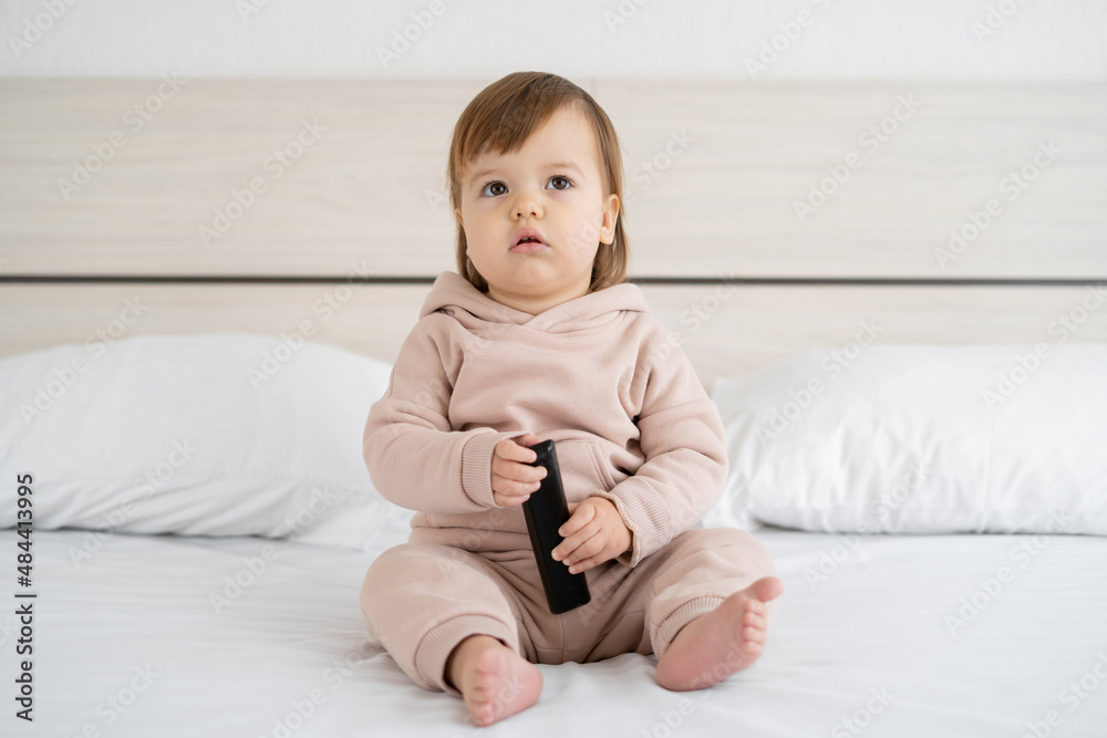 Smiling preschooler girl watching TV while sitting on the bed with a remote control in her hand. Banner. Smiling preschooler girl watching TV while sitting on the bed with a remote control in her hand. Banner.