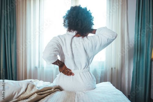 Woman sitting on a bed with back pain. African-american woman having back pain after sleep. Young Black woman suffering from backache at home.