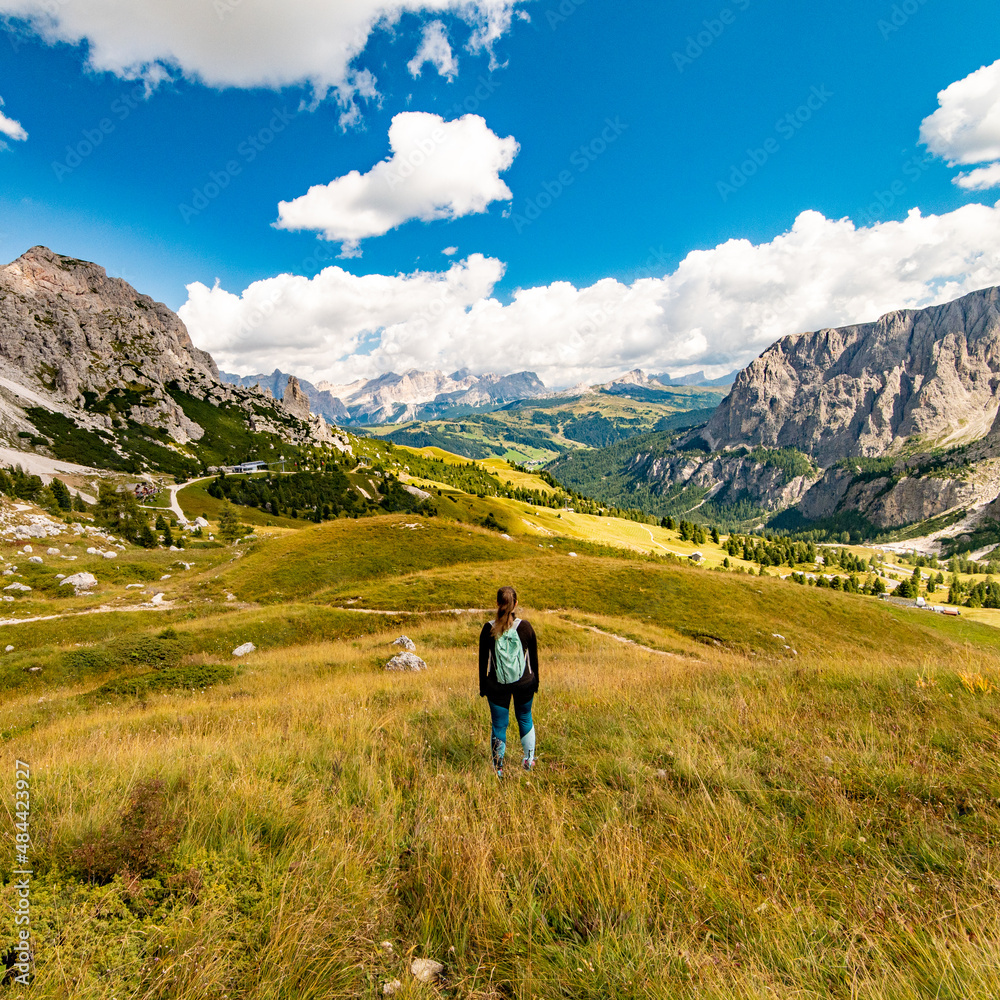 Fototapeta premium Aerial view of active sport woman with turquoise backpack looking at Gardena Pass, Sass da Ciampac, Tor Bornech and Sass dla Luesa, Trentino Alto Adige, Italy. Alpine meadows and Passo Gardena.