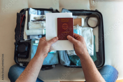 Mens hands putting a face mask and passport in the suitcase prepared to travel, in the new normal, after the coronavirus covid 19 pandemic. High angle view of suitcase with clothes, passport and masks