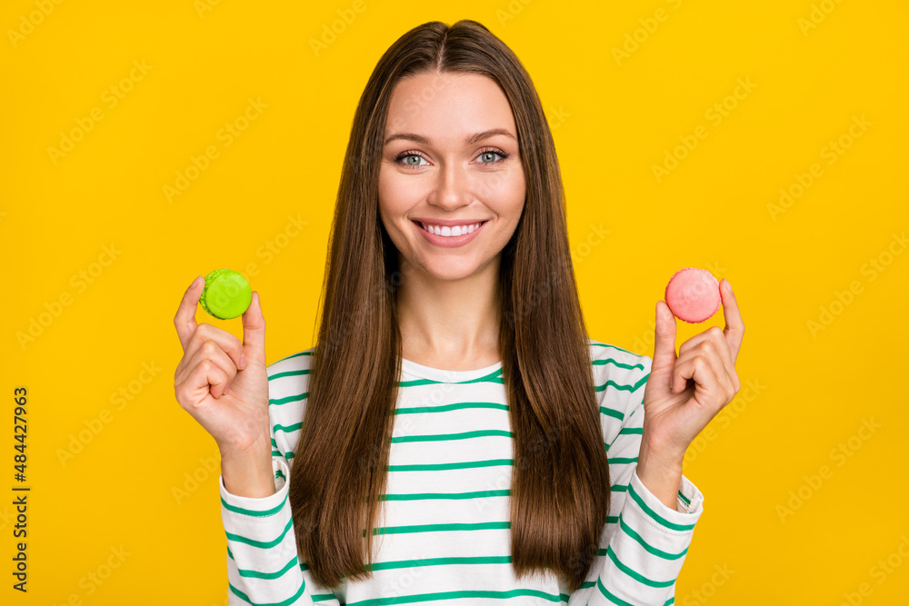 Photo of sweet pretty lady dressed striped shirt choosing green pink cookies isolated yellow color background Photo of sweet pretty lady dressed striped shirt choosing green pink cookies isolated yellow color background