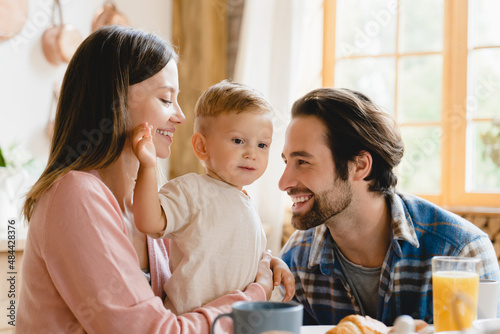 Adoption concept. Caucasian smiling young family of three parents father and mother with small little kid infant toddler new born baby having morning breakfast at home kitchen. IVF