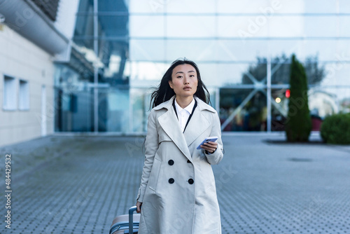 Beautiful female tourist near the airport, a Chinese woman walking with a big suitcase, an Asian woman holding a phone, using the application for booking accommodation and ordering a taxi © Liubomir