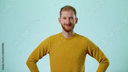 Studio Portrait Of Confident Smiling Bearded Young Caucasian Man With Hands On Hips Against Blue Background. People Lifestyle And Emotions Concept. Looking At Camera In Slow Motion