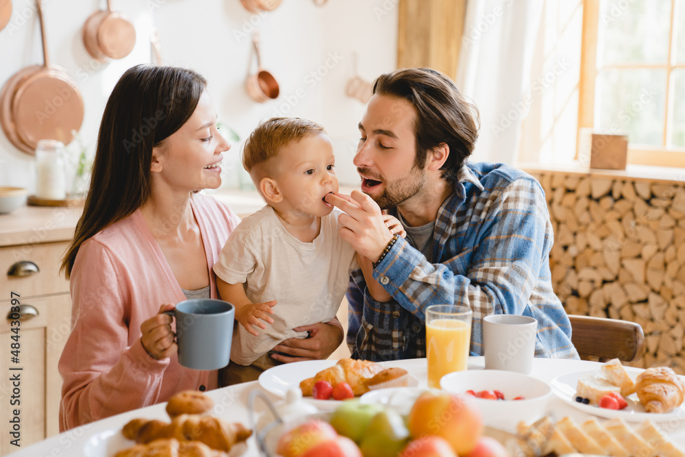Little small caucasian toddler infant new born baby eating berry while dad father feeding child kid. Young family of three having morning breakfast at home kitchen