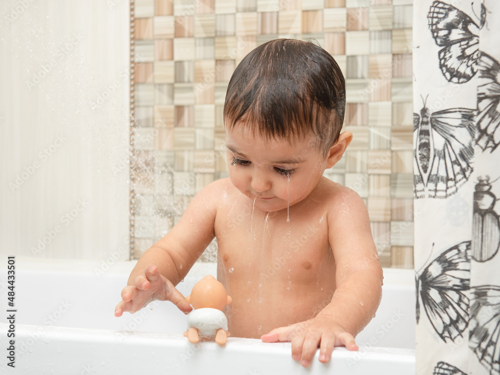 Little boy standing under the stream of water from the shower. The concept of children's hygiene and health care at home. Portrait of a child bathing in the bathroom. Wide open brown eyes. Little boy standing under the stream of water from the shower. The concept of children's hygiene and health care at home. Portrait of a child bathing in the bathroom. Wide open brown eyes.
