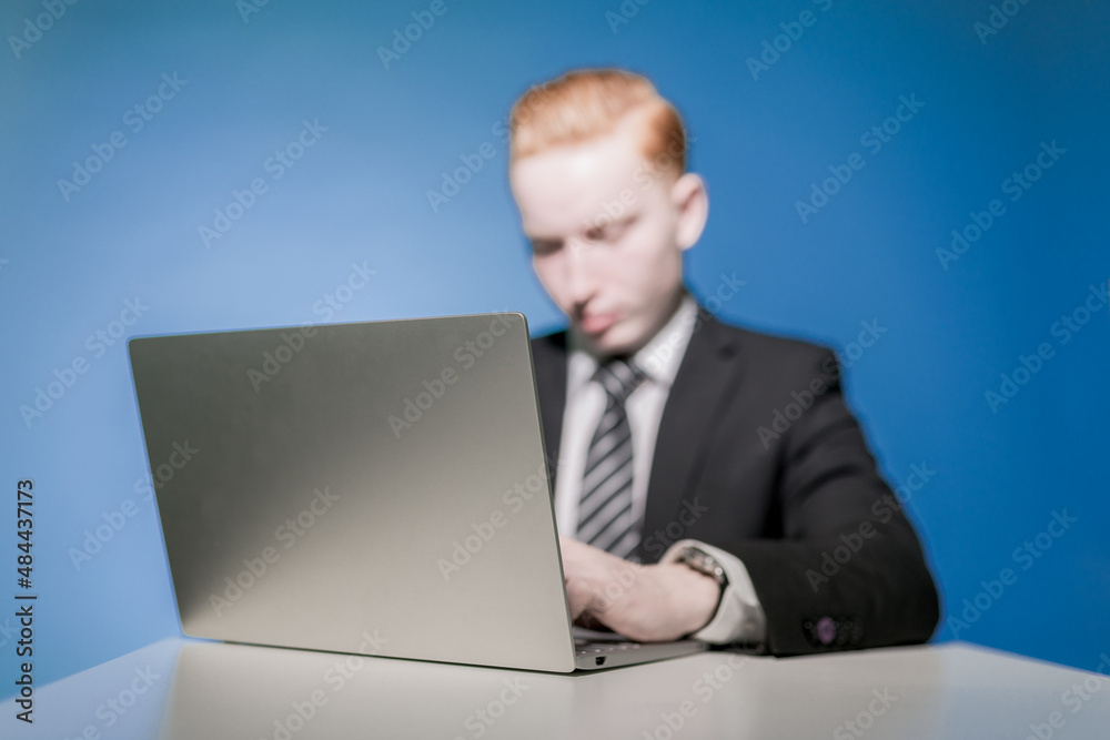 Businessman. Businessman. Portrait of a young red-haired man in a suit, sitting at a table typing text on a laptop on a blue background. Boss. The beginning. Seller. An office worker Businessman. Businessman. Portrait of a young red-haired man in a suit, sitting at a table typing text on a laptop on a blue background. Boss. The beginning. Seller. An office worker