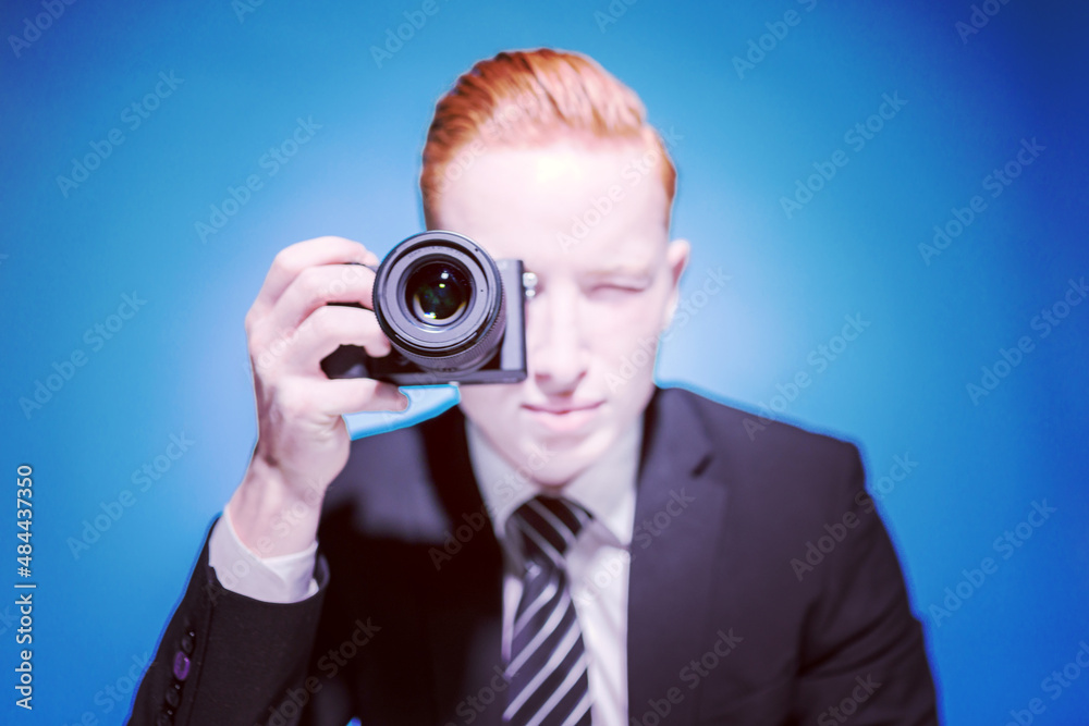 An office worker. Portrait of a young red-haired man in a suit, sitting at a table taking a photo with a camera on a blue background. Atelier. Photographer