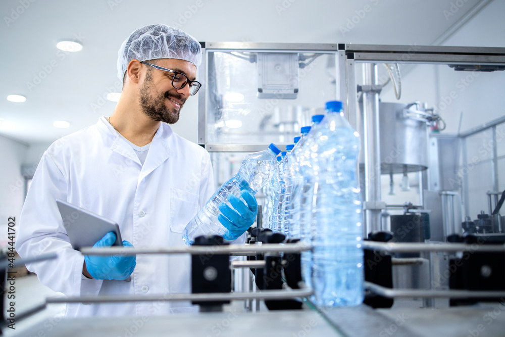 Industrial technologist holding tablet computer and checking production of bottled water in bottling factory on automated machine.