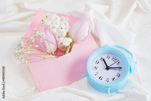 a bouquet of spring flowers in delicate shades, an alarm clock and a pink envelope on the bed, a romantic gift photo