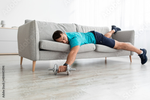 Funny tired young caucasian male lies on sofa with dumbbell in living room interior, rest after training