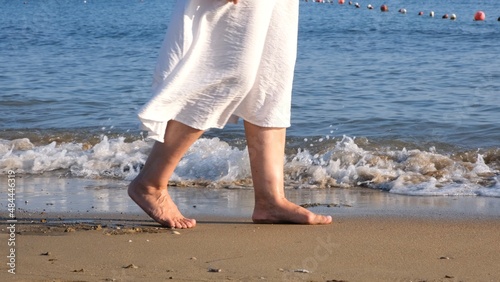 mature female legs are walking along the sandy beach and splashing in the sea on a summer sunny day. Woman walks barefoot on the water close-up