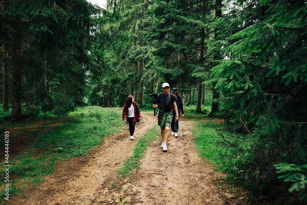Obraz premium Stylish young people walking on trails in a mountain forest and making video content on a hike. Friends walk in nature on a trip in the mountains.