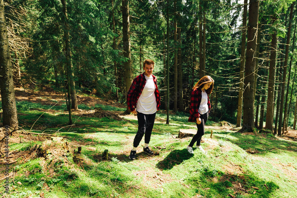 Obraz premium Beautiful man and woman standing in a sunny forest on the ground with moss and resting on a hike. Positive tourists in red shirts walk in the mountain coniferous forest.
