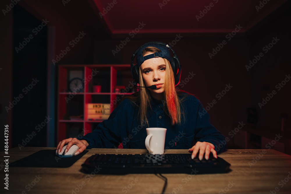 Tired female gamer streaming online games on the computer at home in a cozy room, looking at the camera with a gloomy face and a cup of tea on the table, wearing a headset and cap.