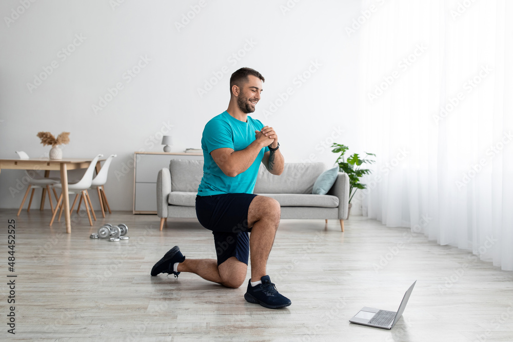 Smiling young muscular caucasian man doing leg exercises and watching online lesson on computer