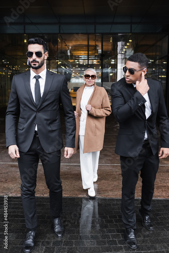 interracial bodyguards in sunglasses escorting stylish businesswoman near hotel building.