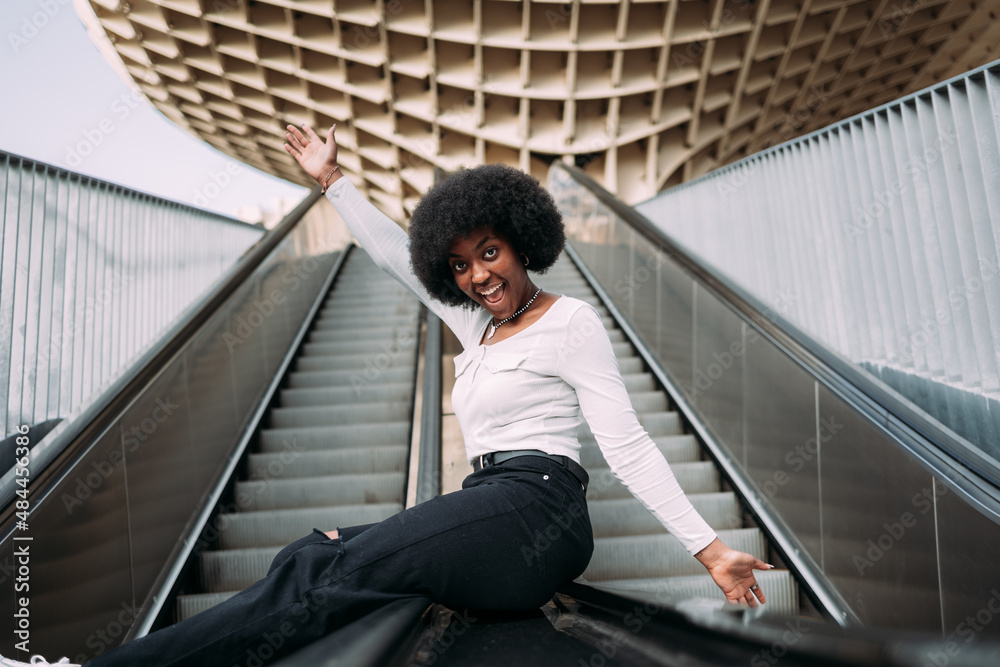 Naklejka premium Smiling and playful young black woman sliding up an escalator railing