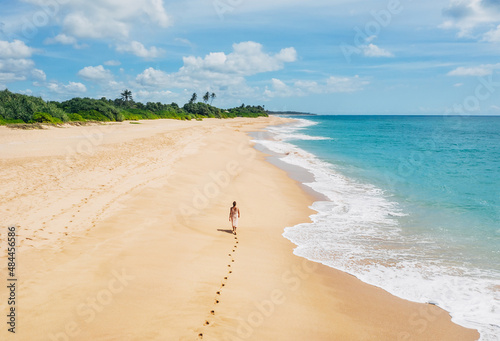 Young female dressed in light summer clothes walking barefoot leaving footprints on the sand on Indian ocean Tangalle lonely coconut trees beach on Sri Lanka island. Aerial top view drone shot.