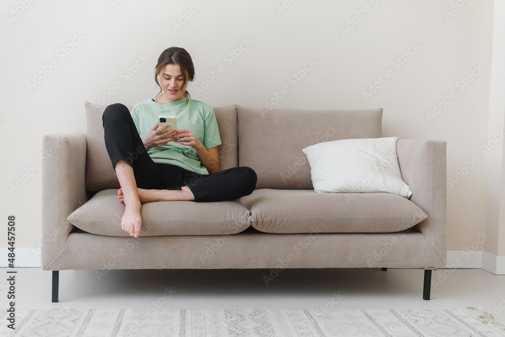 Image of happy optimistic young woman sitting indoors at home and shopping or social media using mobile phone on sofa at home.