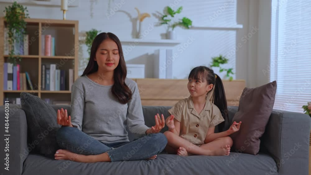 Asian family young happy healthy mom teaching cute little kid daughter meditate together on sofa at home, Mother and small child girl calm to yoga exercise relax together in lotus pose. Asian family young happy healthy mom teaching cute little kid daughter meditate together on sofa at home, Mother and small child girl calm to yoga exercise relax together in lotus pose.