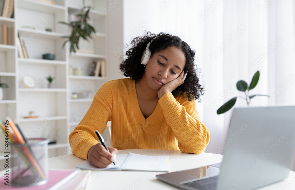 Obraz premium Bored young black female student having dull online lesson on laptop computer, writing down information at home