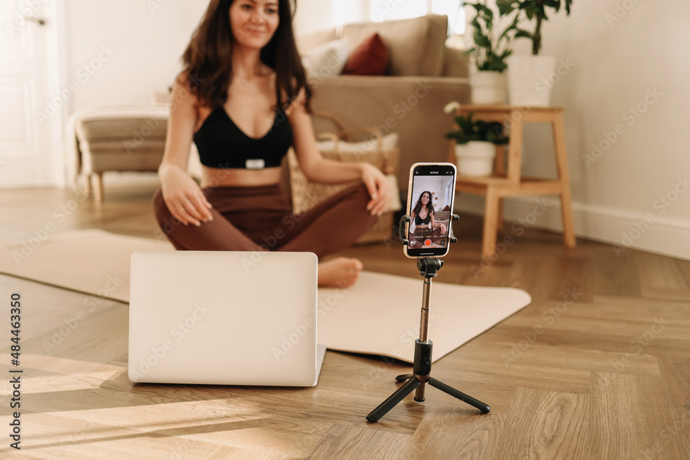 Athletic young millennial woman is engaged in sports training and exercise online in a room at home. A fitness blogger records videos during a workout using a smartphone and laptop. Selective focus Athletic young millennial woman is engaged in sports training and exercise online in a room at home. A fitness blogger records videos during a workout using a smartphone and laptop. Selective focus