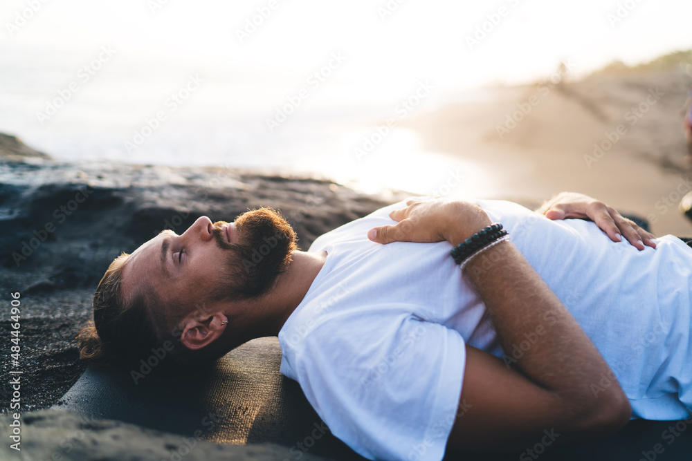 Bearded man with closed eyes lysing and resting during pastime for pranayama breathing in nature environment, calm male yogi feeling mindfulness enlightenment during holistic healing and retreat