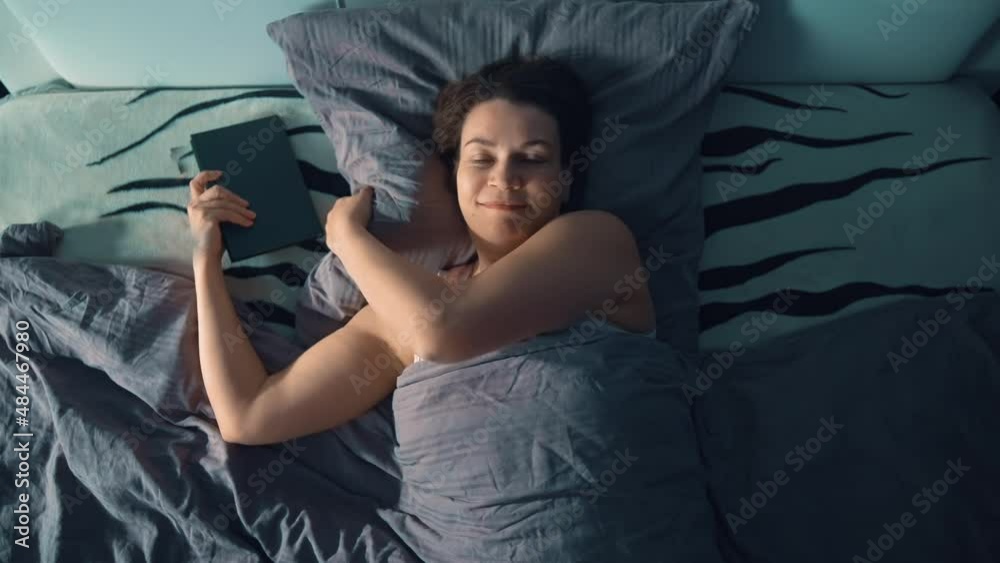 The woman is lying resting alone on the bed. She puts the book she has read under her pillow and looks around in fascination. Evening rest in bed in your free time. Shooting from the top point. The woman is lying resting alone on the bed. She puts the book she has read under her pillow and looks around in fascination. Evening rest in bed in your free time. Shooting from the top point.