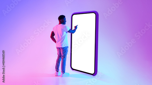 Black guy standing near big cellphone with mockup for your app on screen, interacting with user interface in neon light photo