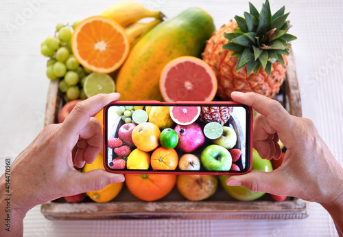 Wooden basket on white tablecloth full of fresh raw fruits. Mature caucasian woman using smart phone to filming recipe to share online internet. Concept of generation X streamer
