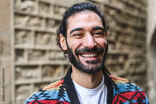 Merry Hispanic man in colorful jacket with beard and ponytail smiling and looking at camera. while standing on blurred background of brick wall on street