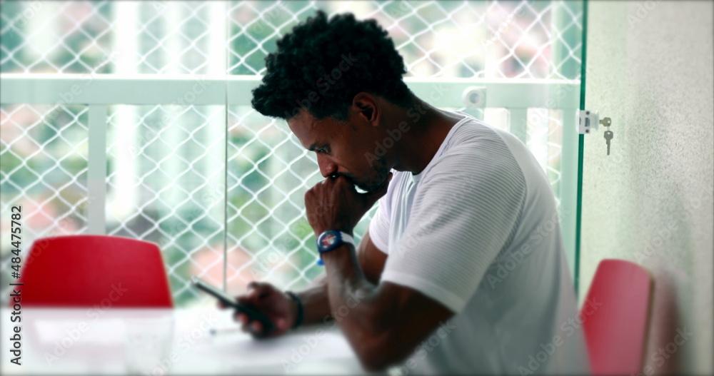 Pensive African man looking at smartphone device at home. Thoughtful black person staring at cellphone screen Pensive African man looking at smartphone device at home. Thoughtful black person staring at cellphone screen