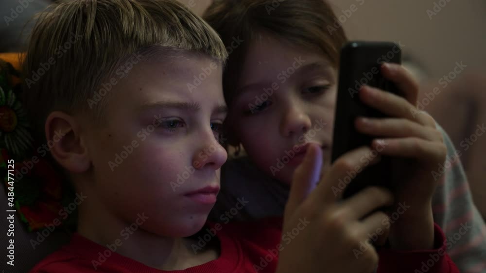 Handsome wistful boy using gadget concept. Serious kid boy is sitting in living room and using smartphone looks at screen, and little gir looks into his phone. Children play phone game app at table Handsome wistful boy using gadget concept. Serious kid boy is sitting in living room and using smartphone looks at screen, and little gir looks into his phone. Children play phone game app at table